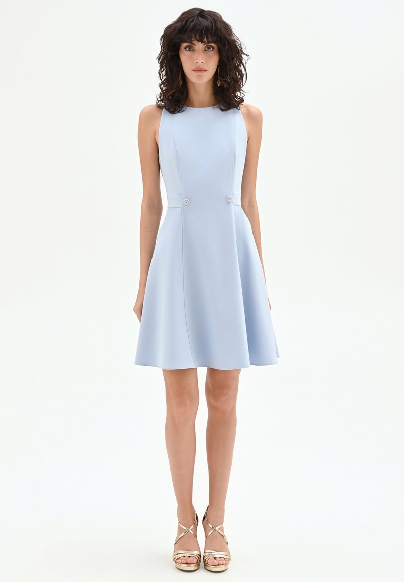 Woman with curly dark hair wearing sleeveless light blue A-line dress and gold strappy high-heeled sandals against white background.