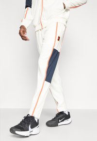 Λευκά αθλητικά παντελόνια με ναυτικό και κοραλλί λεπτομέρειες, που διαθέτουν πλαϊνές ρίγες και λογότυπο. Συνδυασμένα με μαύρα αθλητικά παπούτσια Nike. Λείος υφασμάτινος ιστός.