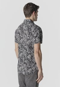 Homme de dos portant une chemise noire à manches courtes avec un motif de feuilles blanches et un jean gris foncé sur un fond uni.
