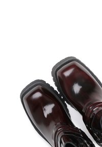 Bottes bordeaux brillantes avec un bout rond, semelle extérieure en caoutchouc noir et œillets en métal pour les lacets, présentant un design texturé sur les côtés.