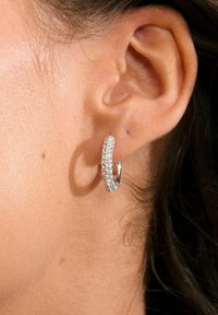 Pendiente de aro plateado adornado con pequeños cristales transparentes, que presenta un diseño liso y redondeado, expuesto en un primer plano de una oreja.