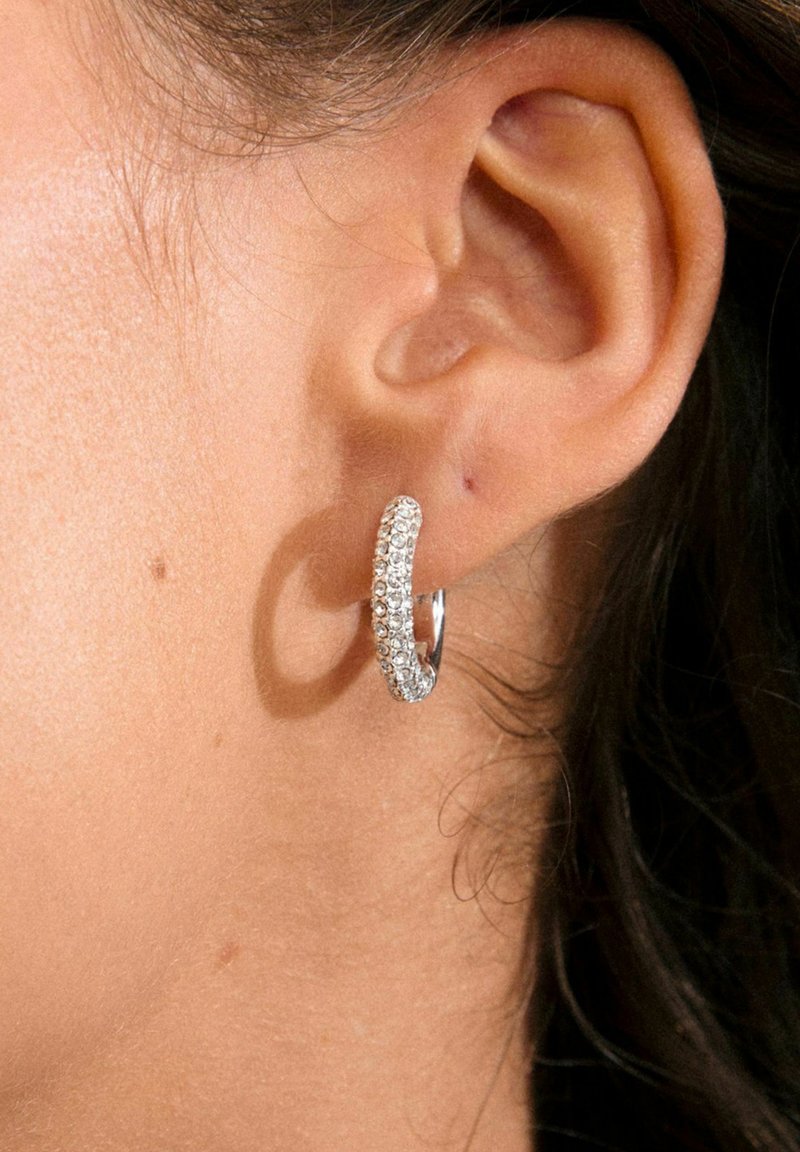 Pendiente de aro plateado adornado con pequeños cristales transparentes, que presenta un diseño liso y redondeado, expuesto en un primer plano de una oreja.
