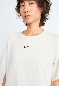Šviesiai smėlinis medvilnės marškinėliai su juodu Nike logotipu ant krūtinės. Trumpomis rankovėmis ir apvaliu iškirpimu, lygaus paviršiaus.