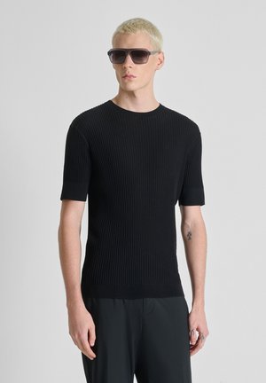 Uomo biondo che indossa occhiali da sole rettangolari scuri, una maglietta nera a maniche corte a coste e pantaloni neri larghi, in piedi davanti a uno sfondo semplice.