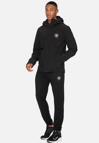 Giacca a windbreaker nera con dettagli del logo. Abbinata a pantaloni jogger neri che mostrano un logo simile. Scarpe sportive nere completano l'outfit.