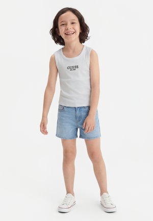 Niño sonriente con cabello rizado, vistiendo una camiseta blanca sin mangas de Guess, pantalones cortos vaqueros claros y zapatillas blancas, posando frente a un fondo blanco.