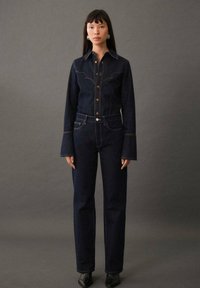 Dunkelblauje Denimjacke mit kontrastierenden Nähten und weit ausgestellten Ärmeln, kombiniert mit hoch-taillierten dunklen Jeans. Enganliegendes Design mit geprägtem Knopfverschluss.