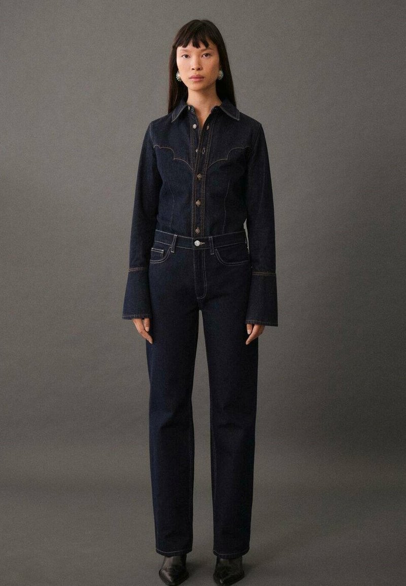 Dunkelblauje Denimjacke mit kontrastierenden Nähten und weit ausgestellten Ärmeln, kombiniert mit hoch-taillierten dunklen Jeans. Enganliegendes Design mit geprägtem Knopfverschluss.