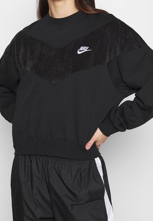 Person wearing a black Nike sweatshirt with a ribbed chevron panel and matching black and white athletic pants, hand on hip.
