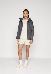 Kvinna står klädd i en mörkgrå huvjacka, vit långärmad tröja, beige shorts med fickor, vita strumpor och beiga sneakers mot en grå bakgrund.