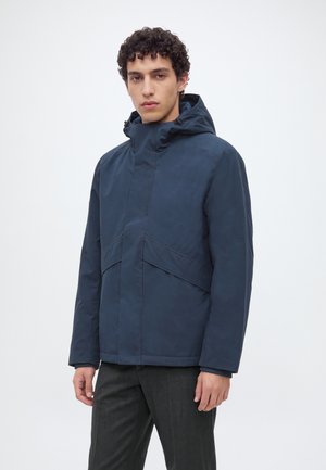 Jeune homme portant une veste d'hiver à capuche bleu foncé et un pantalon sombre, debout devant un fond clair uni.