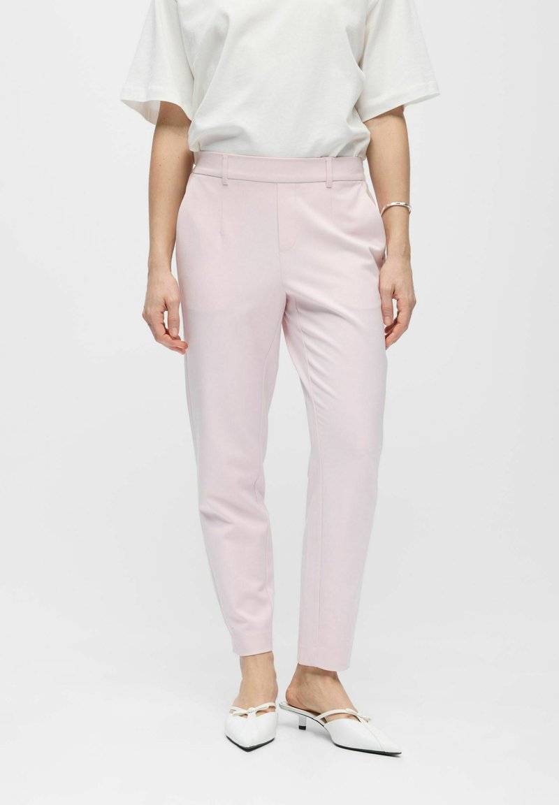 Frau trägt hellrosa maßgeschneiderte Hosen bis zum Knöchel, weißes kurzärmeliges Oberteil, weiße spitz zulaufende Absatzschuhe und ein silbernes Armband, steht.