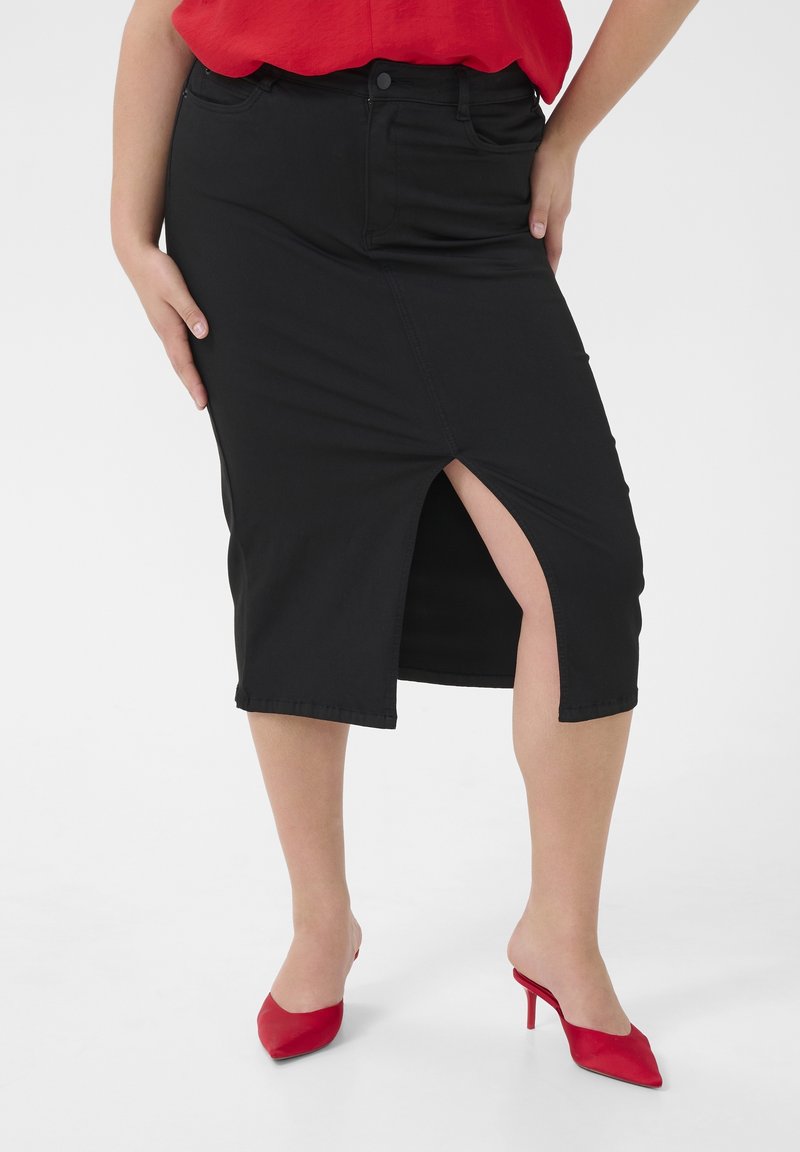 Femme portant une jupe noire mi-longue avec une fente à l'avant, un haut rouge et des chaussures rouges à talons pointus, debout les mains sur les hanches.