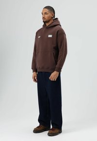 Bruine capuchontrui met een zak aan de voorkant, voorzien van logopatch; gecombineerd met losse donkere spijkerbroek en bruine veterlaarzen.