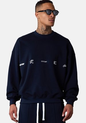 Sweatshirt bleu marine avec un col rond, coupe ample, et logos brodés en blanc sur la poitrine. Tissu doux avec des poignets et un ourlet côtelés.
