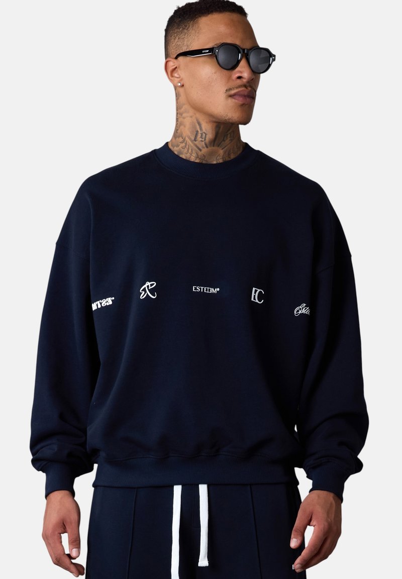 Sweatshirt bleu marine avec un col rond, coupe ample, et logos brodés en blanc sur la poitrine. Tissu doux avec des poignets et un ourlet côtelés.