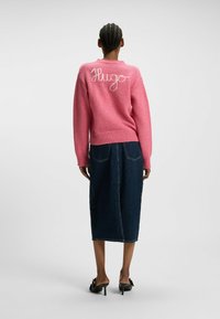 Rosa Strickpullover mit weißer "Hugo" Stickerei auf dem Rücken, kombiniert mit einem langen dunklen Jeansrock und schwarzen, hohen Schuhen.