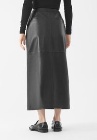 Vrouw in een zwarte lange leren rok, zwarte loafers en een zwart langslavend top, staand tegen een effen witte achtergrond.