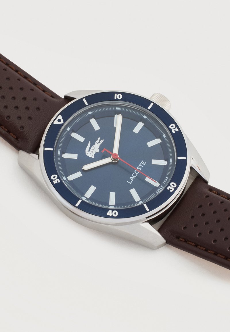 Zilveren horloge met een blauwe wijzerplaat, witte uurmarkeringen, rode secondenwijzer, Lacoste-logo en geperforeerde bruine leren band op een witte achtergrond.