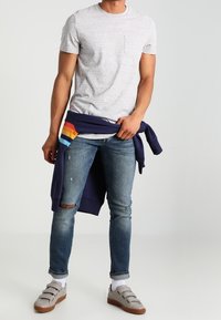Camiseta gris de cuello redondo, jeans azules desgastados, sudadera navy atada a la cintura, calcetines blancos y zapatos slip-on grises con suelas marrones.