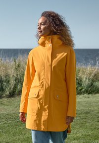 Gul vattentät jacka med hög krage, tryckknappar och två framfickor. Mjuk textur, knälång design, bärs över blå jeans.