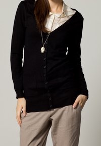 Black knit cardigan with a deep V-neck and six front buttons, paired with a light beige shirt, worn over relaxed beige trousers.