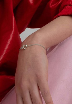 Silverfärgad kedjebr bracelet med ett hjärtformat hänge med en texturerad design, uppvisad på en hand mot en rosa bakgrund.