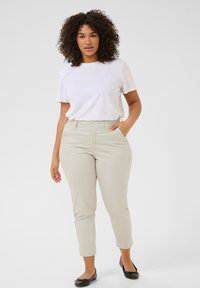 Pantaloni beige chiari con una vestibilità slim, tasche anteriori e chiusura a un bottone, abbinati a una maglietta bianca semplice e ballerine nere.