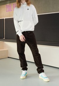 Bílá mikina s kapucí a logem, zkombinovaná s tmavě hnědými kordovědnými kalhotami a bílými teniskami se světle modrými pruhy. Minimalistický design, volný střih.