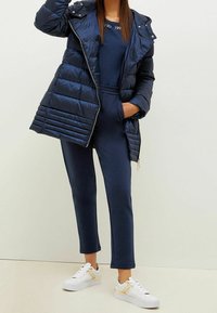 Tmavě modrá prošívaná bunda s vodorovným stehem, doplněná o sladěnou soupravu a bílé tenisky se zlatými detaily. Hladká textura, zapínání na zip.