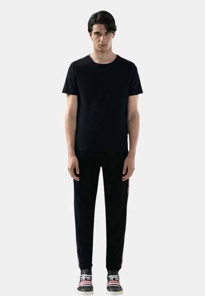 Uomo in piedi, rivolto verso la fotocamera, che indossa una maglietta nera semplice, pantaloni neri con strisce laterali e scarpe da ginnastica nere con dettagli rossi e bianchi.