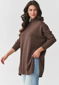 Brązowy sweter dziany z wysokim dekoltem, długimi rękawami, ściągaczami na mankietach i bocznymi rozcięciami, w połączeniu z jasnoniebieskimi dżinsami.