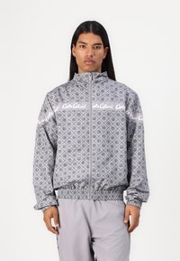 Grijze zip-up jas met een overal geometrisch patroon en logo-accenten. Voorzien van elastische manchetten en tailleband. Glad, lichtgewicht materiaal.