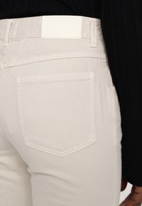 Gros plan sur la poche arrière et la taille d’un jean en denim écru porté avec un haut noir à manches longues côtelé.