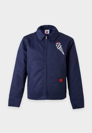 Un mannequin masculin se tient devant un fond uni, vêtu d’une casquette beige, d’une veste bleu marine zippée avec des écussons, d’un pantalon beige et de baskets noires.