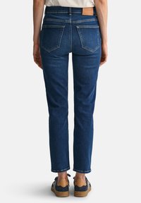 Dunkelblaue Jeans mit geradem Schnitt, hoher Taille und Gesäßtaschen. Kombiniert mit marineblauen und beigen Schuhen sowie einem beigen Hemd.