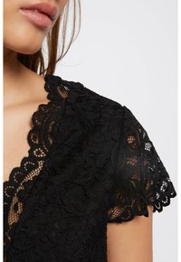 Top in pizzo nero con bordi scallop, un motivo floreale e maniche corte. La texture è complessa e consente una certa trasparenza.