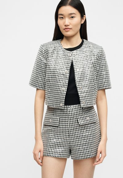 Jeune femme portant une veste à manches courtes à motif pied-de-poule noir et blanc et un short taille haute assorti, debout devant un fond uni.