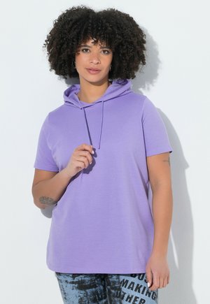 Kobieta o kręconych włosach nosi krótkim rękawem lawendową bluzę z kapturem i wzorzyste dżinsy, stoi na białym tle.
