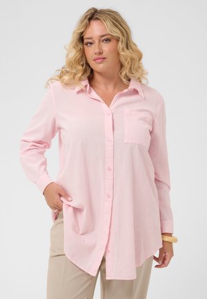 Femme blonde aux cheveux bouclés portant une chemise rose clair à boutons et un pantalon beige, debout avec une main dans la poche.
