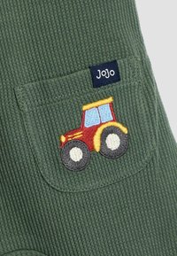 Grüner gerippter Stoff mit einer kleinen Tasche, die einen bunten gestickten Traktor und ein dunkelblaues Etikett mit der Aufschrift "JoJo" zeigt.