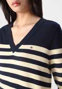 Granatowo-kremowy sweter w paski z dekoltem w serek, wykonany z dzianiny, z małym haftowanym logo na lewej piersi. Miękka faktura i dopasowany fason.