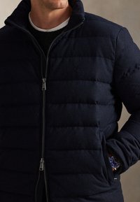 Giacca imbottita trapuntata blu navy con collo alto e cerniera argento. Presenta tasche laterali e un logo sul petto. Tessuto strutturato.