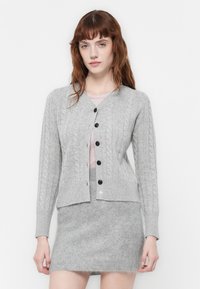 Donna che indossa un cardigan grigio chiaro a trecce con bottoni scuri su una maglietta rosa chiaro e una gonna grigia pelosa abbinata, in piedi contro uno sfondo bianco.