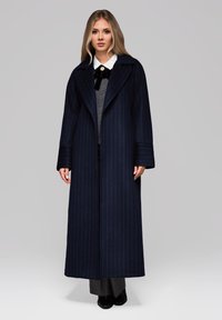Abrigo largo de rayas marinas con solapas anchas, bolsillos laterales y un corte relajado. Superpuesto a un suéter gris y pantalones negros. Acento de lazo de satén.