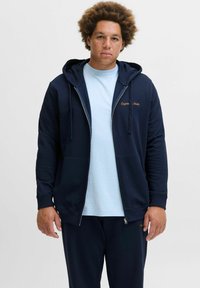 Navy zip-up hoodie van een katoenmix, met een kangoeroe-pocket en geborduurd "Origin Studio" logo. Gedragen over een lichtblauw shirt.