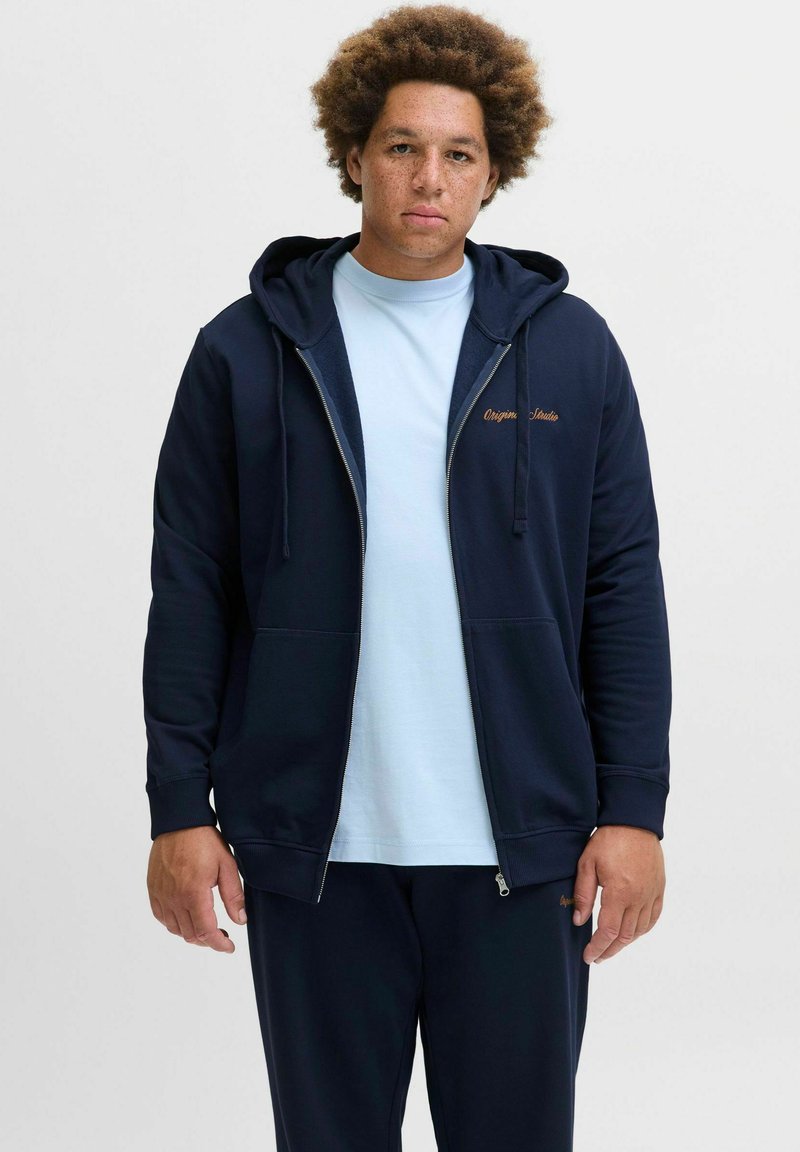 Tamsiai mėlynas zip-up gobtuvas, pagamintas iš medvilnės mišinio, su kengūros kišenę ir siuvinėtu "Origin Studio" logotipu. Nešiojamas ant šviesiai mėlynos marškinėlės.