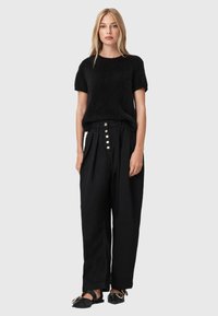 Jersey negro de punto con mangas cortas combinado con pantalones negros de cintura alta que cuentan con una fila de botones plateados. Sandalias planas minimalistas.