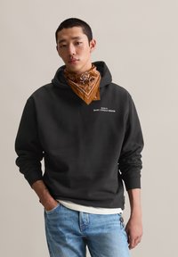 Zwarte hoodie met een kangoeroezak en geribde manchetten, met witte tekst. Draagt over een wit shirt en gecombineerd met lichtblauwe jeans. Brown bandana.