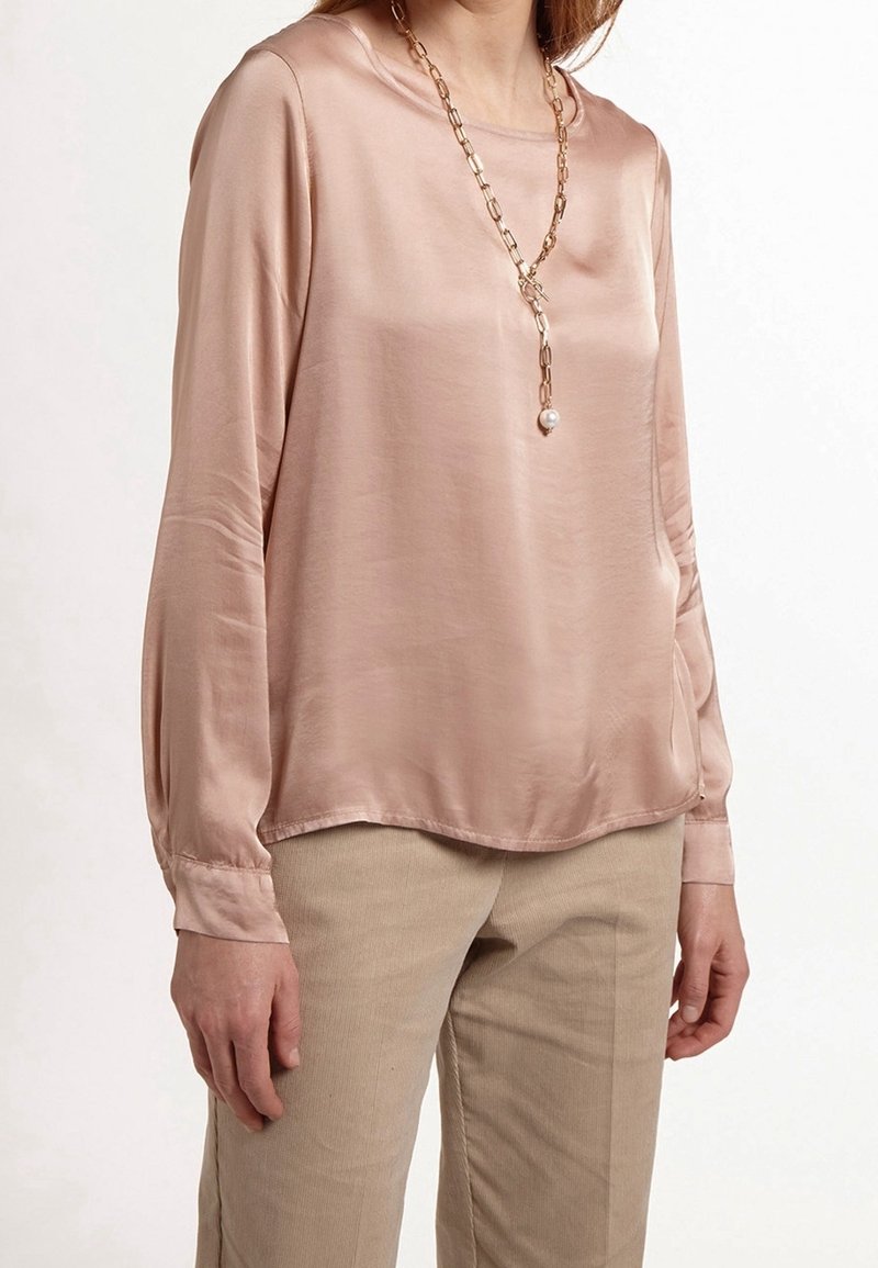 Blusa de satén rosa claro de manga larga con escote redondeado y dobladillo ligeramente curvado, combinada con pantalones beige. El collar presenta un acento de perla.